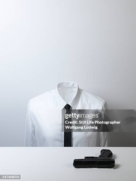 a headless body in a white shirt with a black tie sits in front of a black gun on a plain white desk - headless stock-fotos und bilder