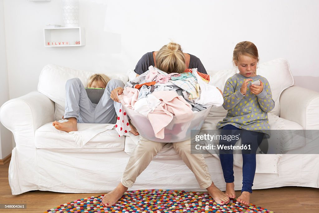 Exhausted mother with laundry basket on couch with children using digital tablet and cell phone