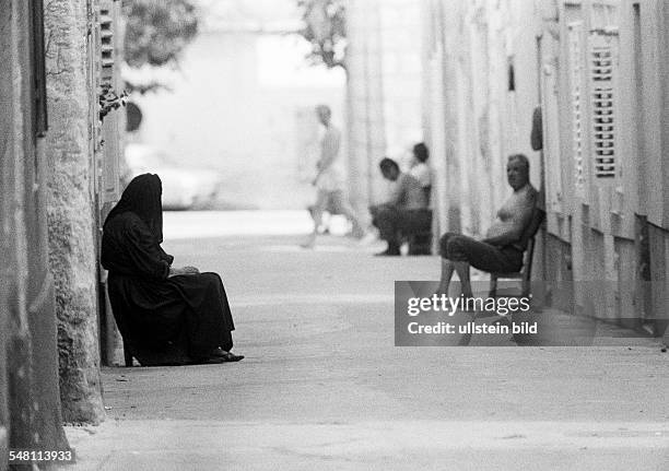 People, midday break, siesta, view into a narrow alley of a village, during the hot noonday people sit in the shadow of their houses holding a...