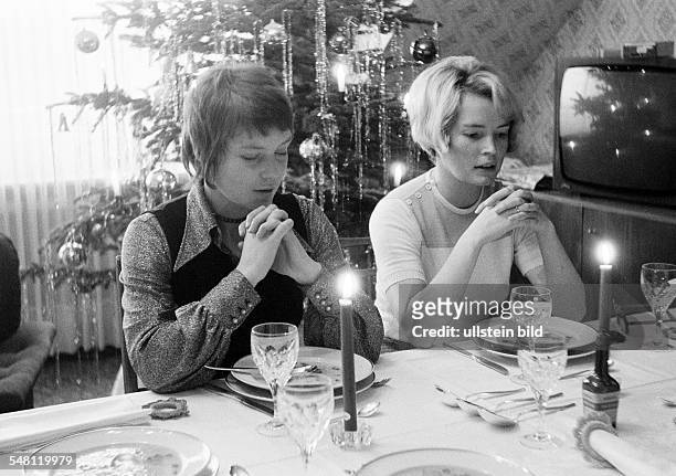 People, food, festivity, Christmas, two young women sit at the well-laid table in the living room saying grace, in the background a Christmas tree,...