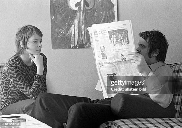 People, young couple sitting in the living room, the man reads a newspaper and ignores the woman, the woman looks very disappointed, aged 20 to 30...