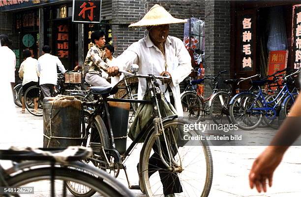 Mann mit Strohhut und Zigarette schiebt sein Fahrrad durch die Strassen von Qufu - Juni 1996