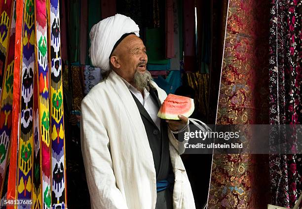 China Xinjiang Uyghur Kashgar or Kashi - Uigur man with turban at sunday market is eating a water melone