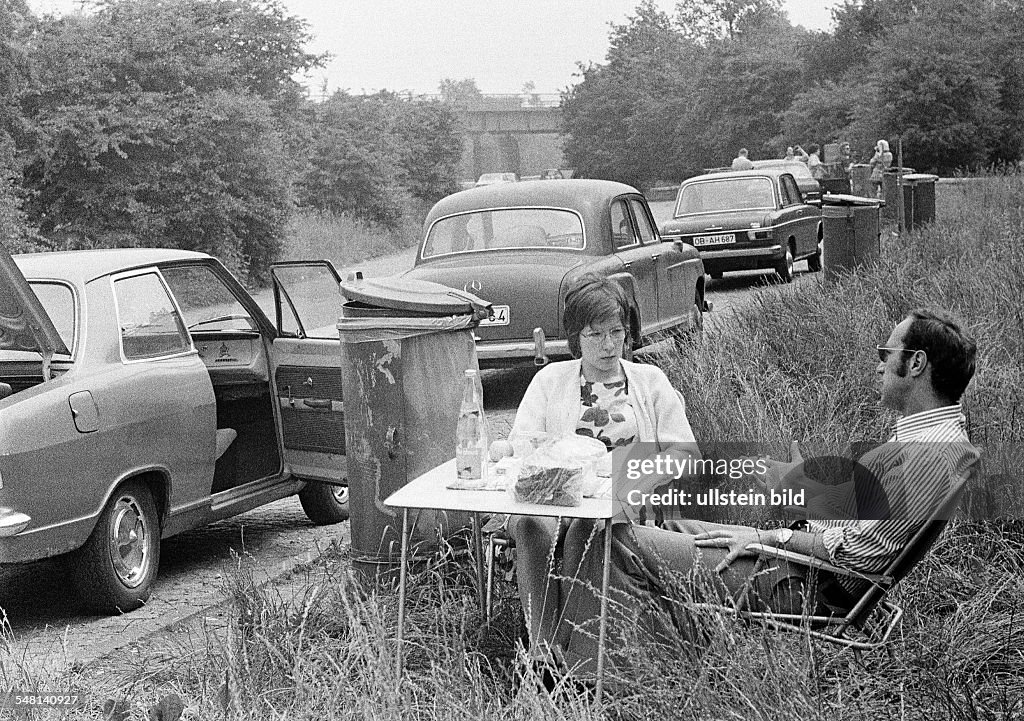 People, freetime, young couple takes a rest from motoring in a rest area, aged 25 to 35 years, picnic with folding chairs and a folding table, in the background parking cars and a dustbin - 30.06.1973