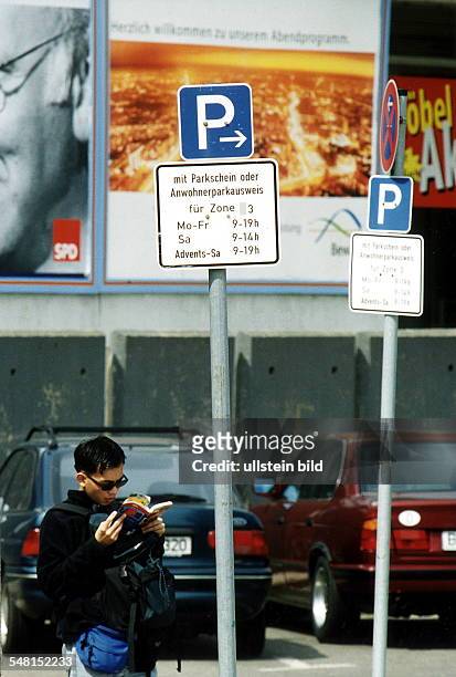 Hinweisschild auf einem gebührenpflichtigen Parkplatz in Berlin Mitte; vor dem Schild ein asiatischer Tourist mit Rucksack, der einen Reiseführer...
