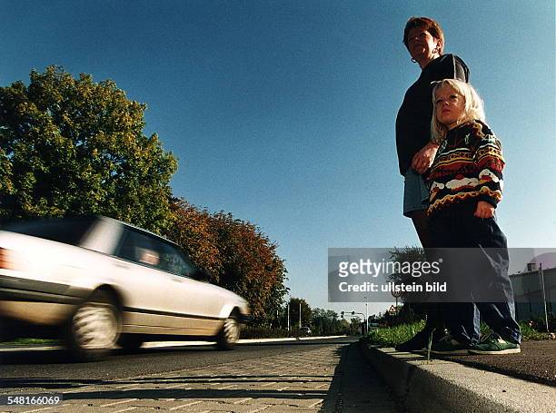 Kinder im Straßenverkehr: ein kleines Mädchen wartet an der Hand ihrer Mutter ein vorbeifahrendes Auto ab, um die Straße überqueren zu können. -...