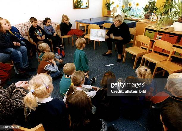 Kinder sitzen im Halbkreis um eine Erzieherin, die ihnen ein Bild in einem Buch zeigt - Oktober 1997