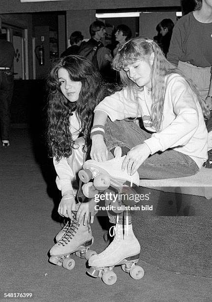 People, two young girls sit side by side and put on their rollerblades, jeans trousers, pulli, aged 15 to 18 years