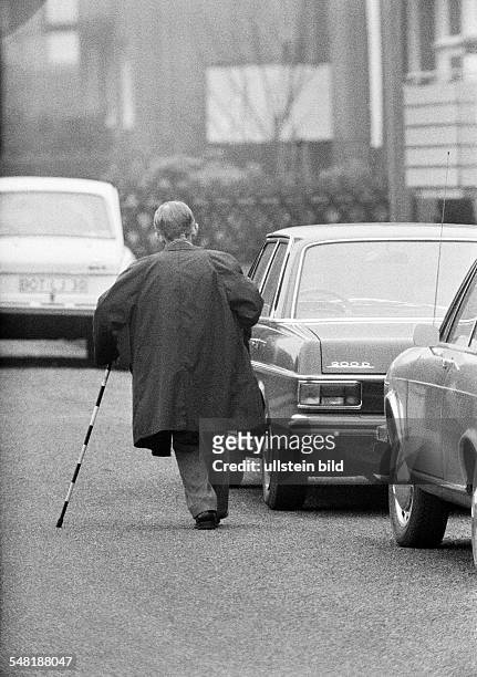 People, older people, older man with a blindmans stick walks on the street and passes some cars, aged 60 to 70 years -