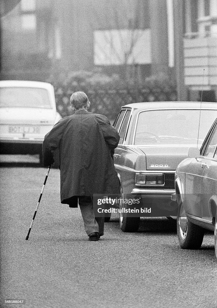 People, older people, older man with a blindmans stick walks on the street and passes some cars, aged 60 to 70 years - 28.01.1973