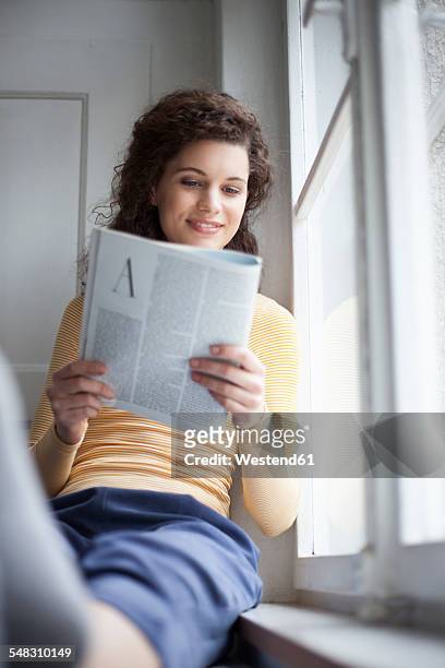 smiling young woman reading magazine at the window - reading magazine stock pictures, royalty-free photos & images