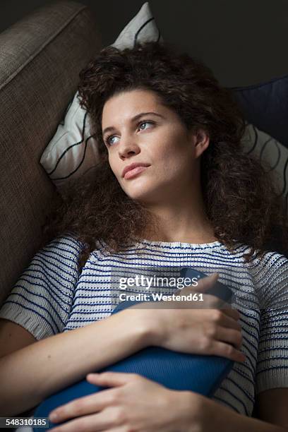 serious young woman lying on sofa with hot water bottle - wärmflasche stock-fotos und bilder
