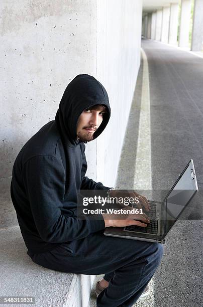 portrait of hacker with laptop sitting in an underground car park - ominous computer stock pictures, royalty-free photos & images