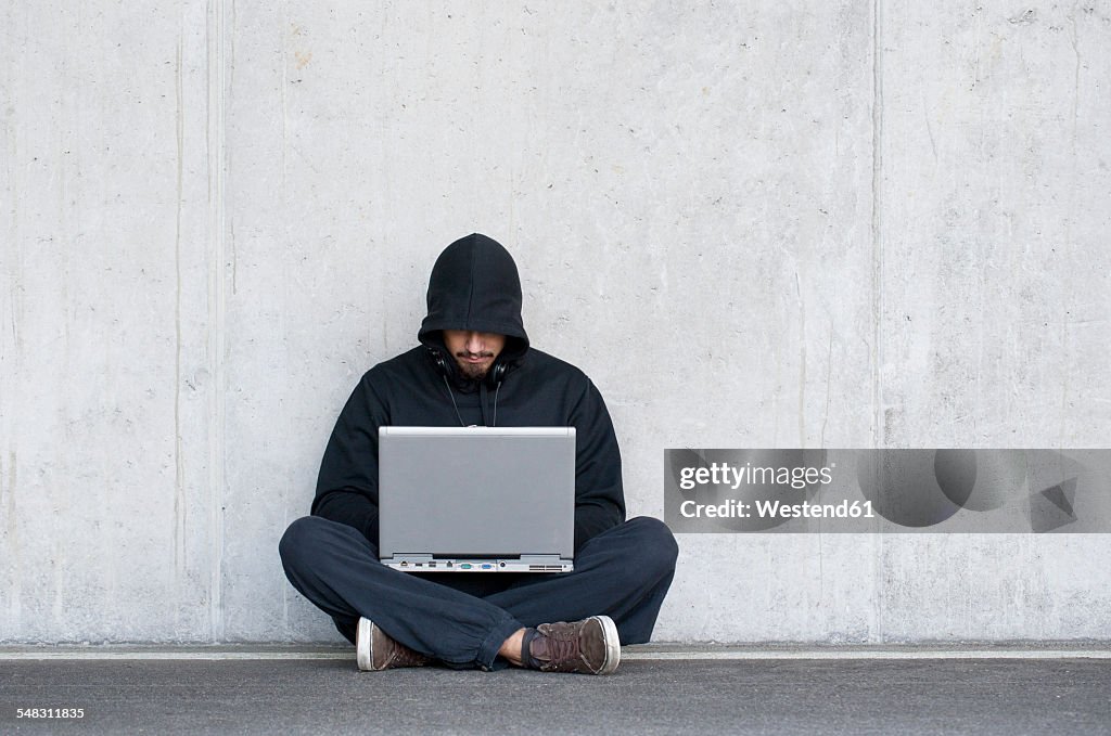 Hacker with laptop sitting in front of concrete wall