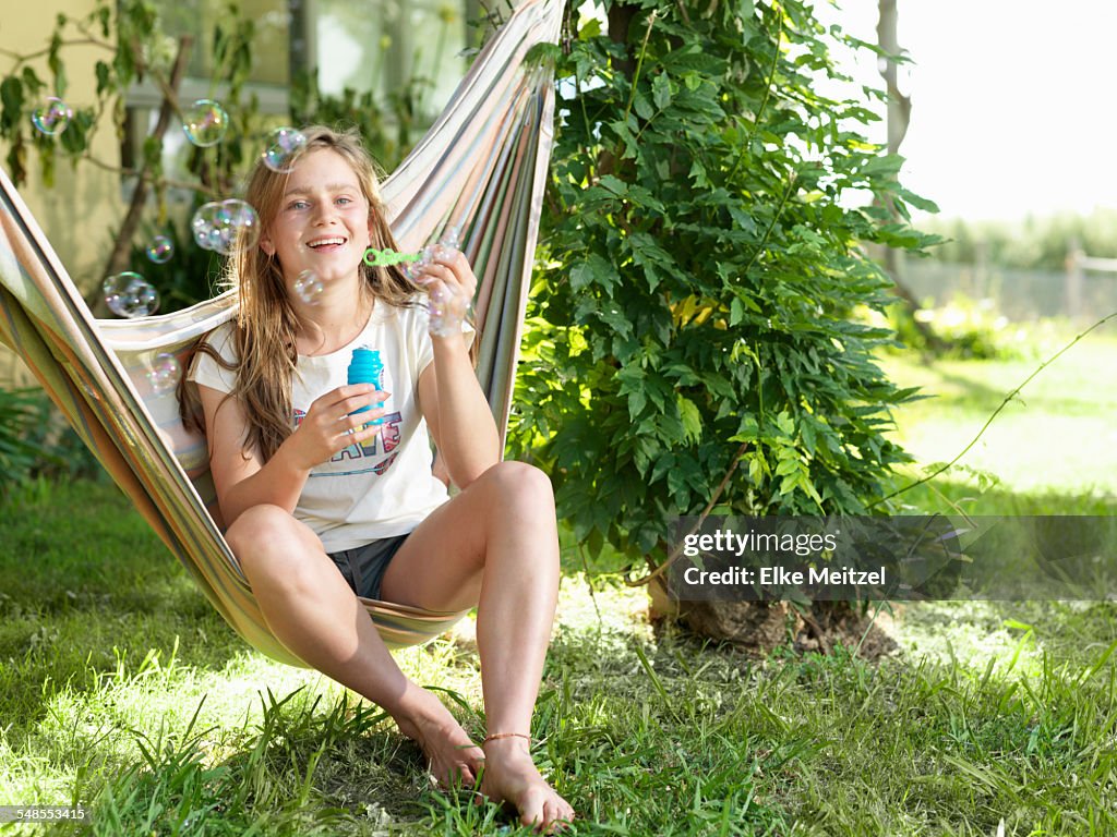 Teenage girl sitting on hammock, blowing bubbles