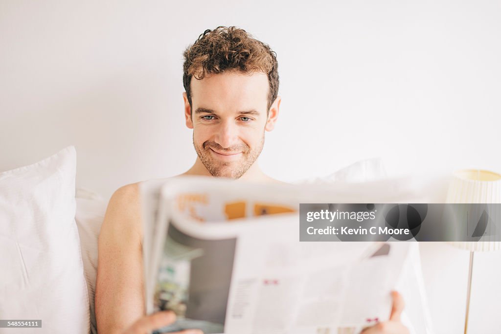 Young man lying in bed smirking whilst reading newspaper