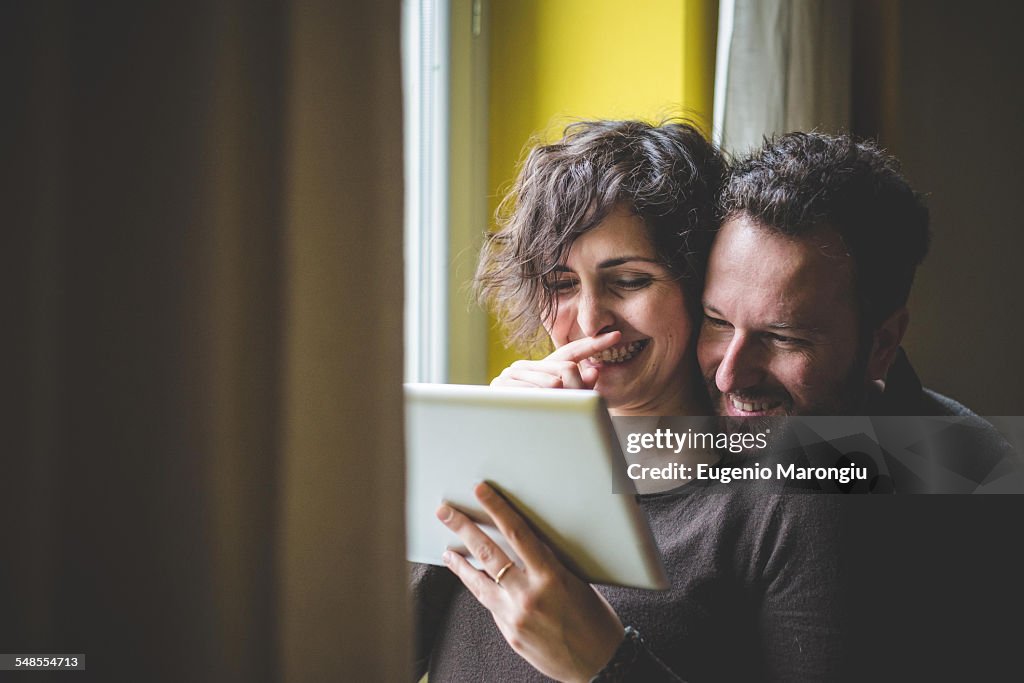 Couple standing beside window, using digital tablet