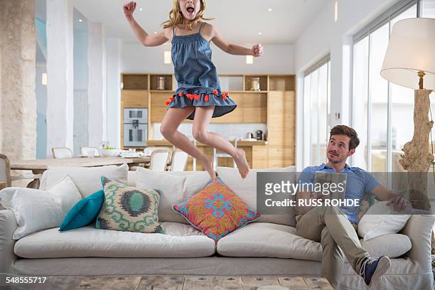 girl jumping mid air from living room sofa whilst father uses digital tablet - jump on sofa stock-fotos und bilder