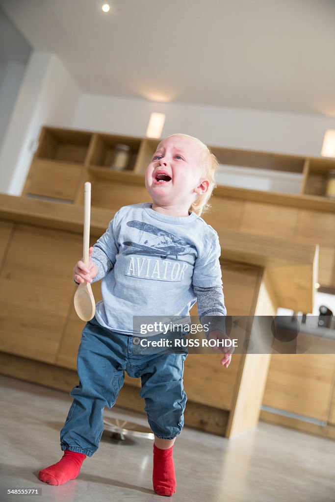 Male toddler holding wooden spoon and crying in dining room