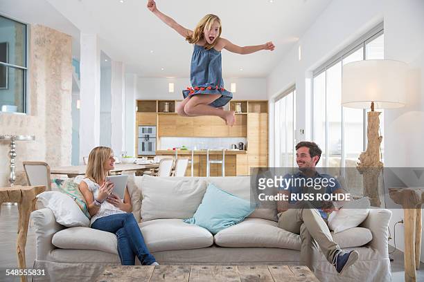 girl jumping mid air from living room sofa whilst parents use digital tablet - gruppe springen ipad stock-fotos und bilder