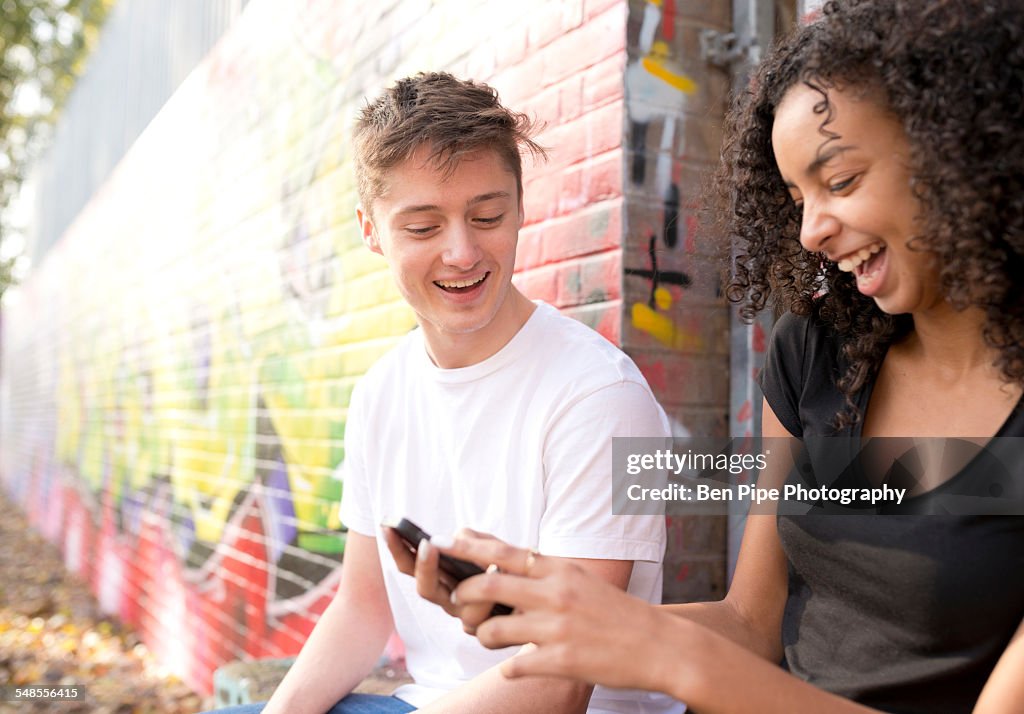 Teenage couple using smartphone against wall with graffiti