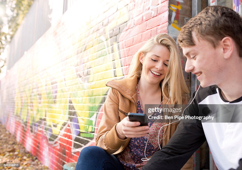 Teenage couple listening to mp3 player against wall with graffiti