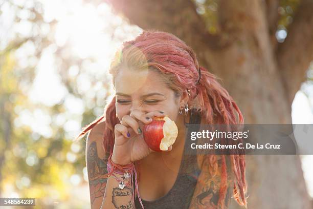 young woman with pink dreadlocks giggling whilst eating apple in park - woman eating fruit stock-fotos und bilder