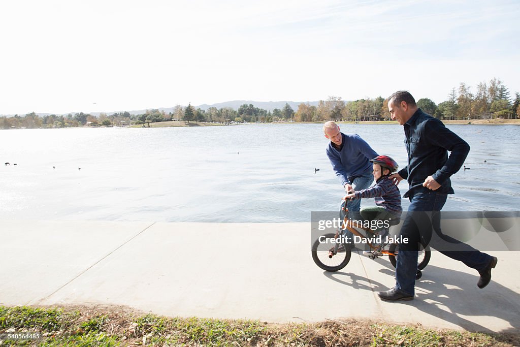 Male couple running next to son on bicycle in park