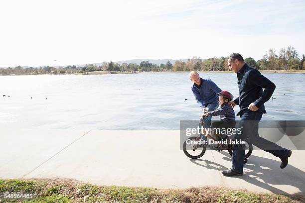 male couple running next to son on bicycle in park - photograph 51 play stockfoto's en -beelden