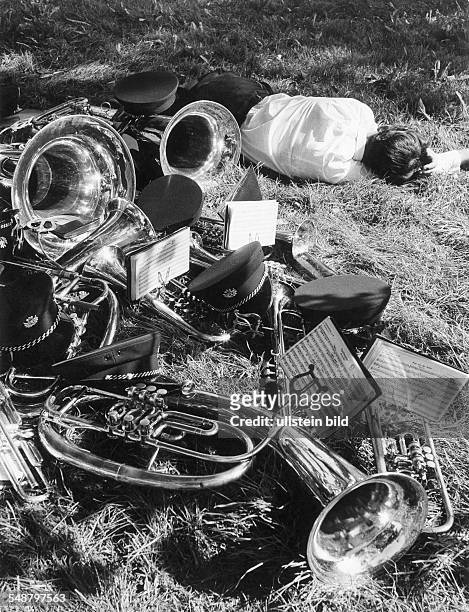 Blechblasinstrumente und Muetzen einer Kapelle liegen im Gras - daneben ein Musiker - um 1965 Aufnahme: Siegfried Lauterwasser