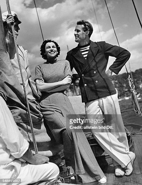 Fashion - Summer Fashion: Woman and man on a sailboat, she wearing elegant jersey pants and a hand-knit sweater with turtle neck collar, he wearing a...