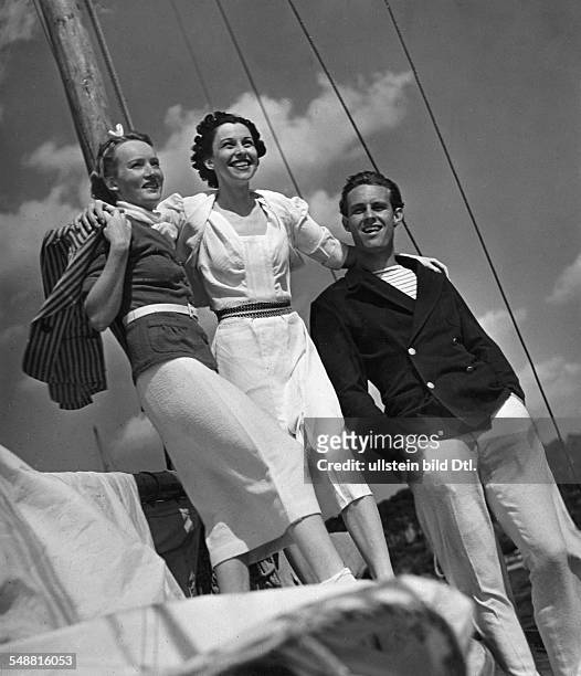 Fashion - summer fashion: two women and a man on a sailboat, the woman on the left wearing a suit in white and red terry cloth and a red-white blazer...