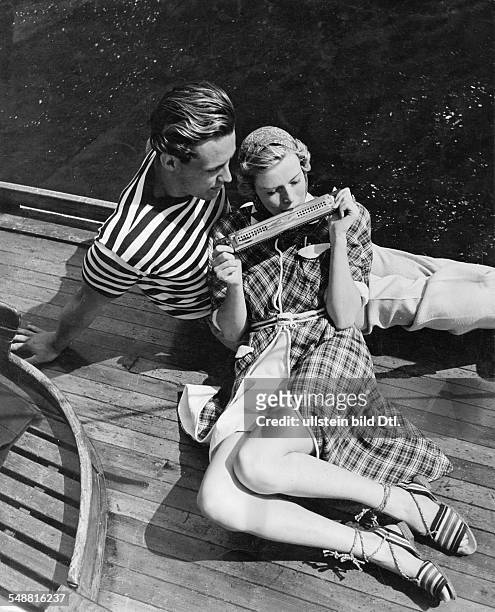 Fashion - summer fashion: woman and man playing harmonica on a sailboat, she wearing a checked linen bathrobe lined with white terry cloth, he...