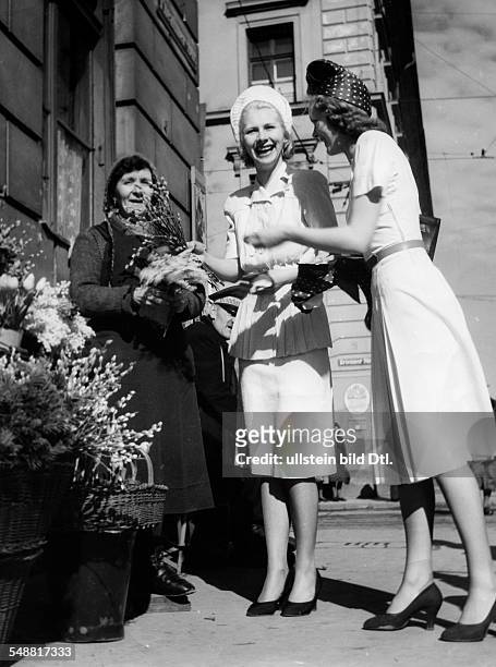 Two young ladies, dressed by the Master School for Fashion in Munich, buying catkins at a florist - around 1941 - Photographer: Hedda Walther Vintage...