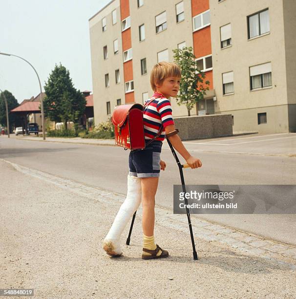 Boy with a leg in plaster and walking frames is crossing a road - 1970s