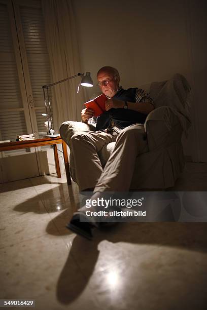 Deutschland Aelterer Mann beim lesen eines Buches im Wohnzimmer bei Kunstlicht