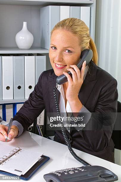 Eine freundliche Frau telefoniert an ihrem Schreibtisch im Büro und notiert Termine im Kalender