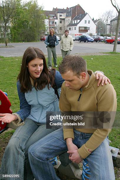 Germany, : Free time.- A young woman and a young man sitting on a playground. She comfortly put an arm around his shoulders while they are talking....