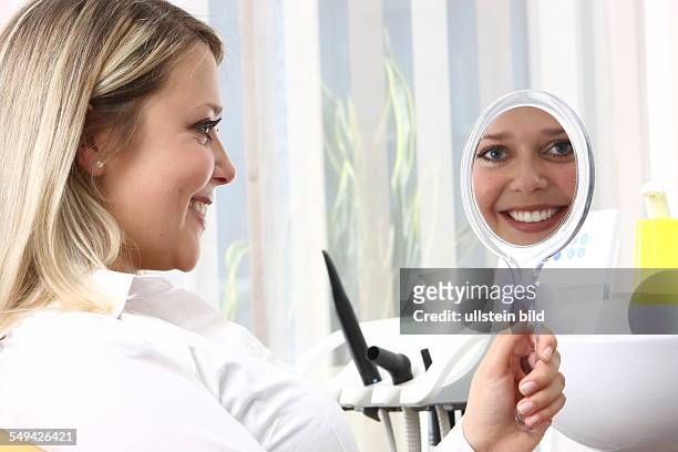 Germany: At a dentist.- A young woman is sitting in a dentist's chair while she is looking in a hand mirror.