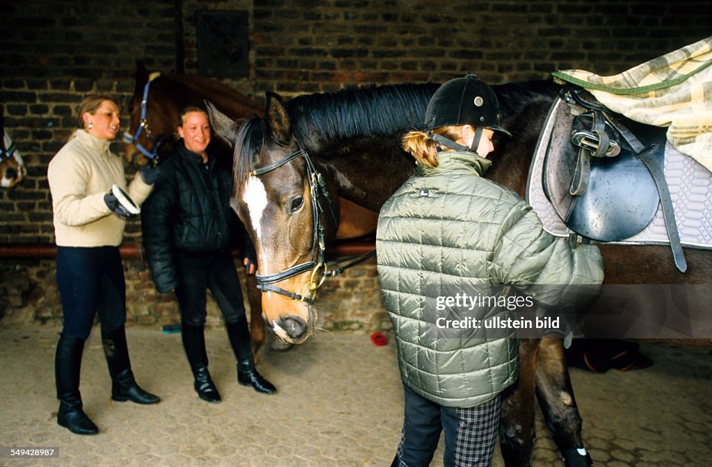DEU, Germany: Free time.- Two young women on a stud farm; they are saddling and curriing the horses.