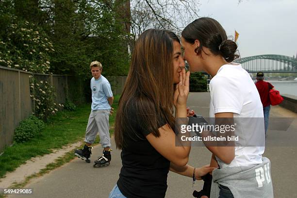 Germany, : Free time.- Two young women skating; they are watched by a boy and whisper.