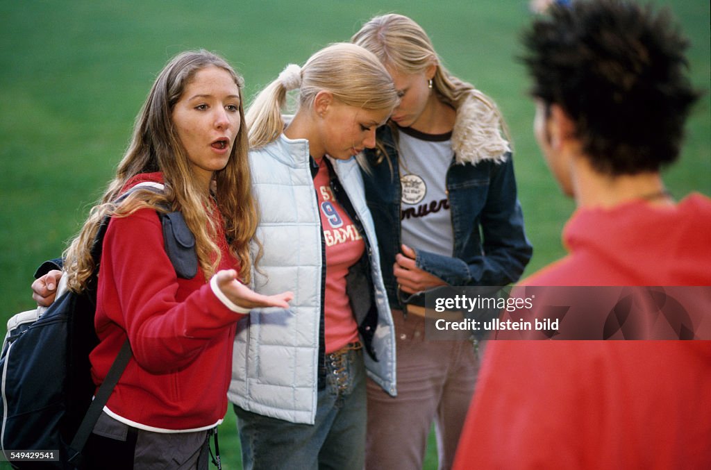 DEU, Germany: Free time.- Four youth on a meadow; the girls embracing each other while one of them is talking to a boy.