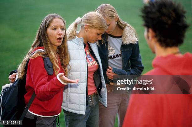 Germany: Free time.- Four youth on a meadow; the girls embracing each other while one of them is talking to a boy.