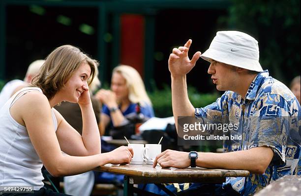 Germany: Free time.- Two young people in a cafe; they are sitting on a bench, talking and drinking coffee.