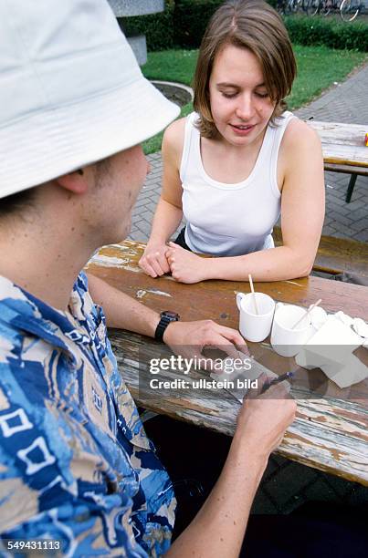 Germany: Free time.- Two young persons in a cafe; the man is writing something on a piece of paper, the woman is watching him.