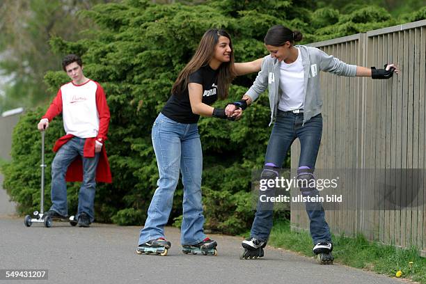 Germany, : Free time.- Two young women trying to skate; a boy is standing in the background watching them.