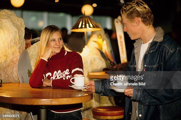 Germany: Free time.- Two young persons at a table; the boy is serving two cups of hot chocolate.