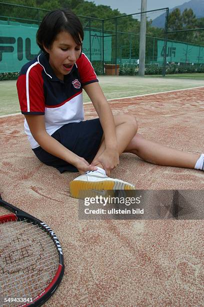 Germany, : On the tennis court.- A young woman is holding her foot because she twisted her ankle.