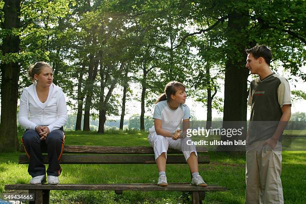 Germany, : Free time.- Three young persons talking in a park; the girls are sitting on a wooden bench.
