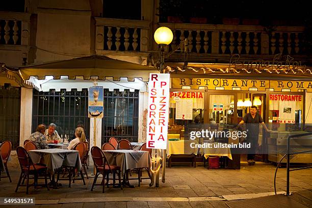 Ristorante Roma Cannaregio 30121 Venezia, Der Kellner steht am Abend vor seinem Restaurant und schaut auf die drei Gäste an einem Tisch.